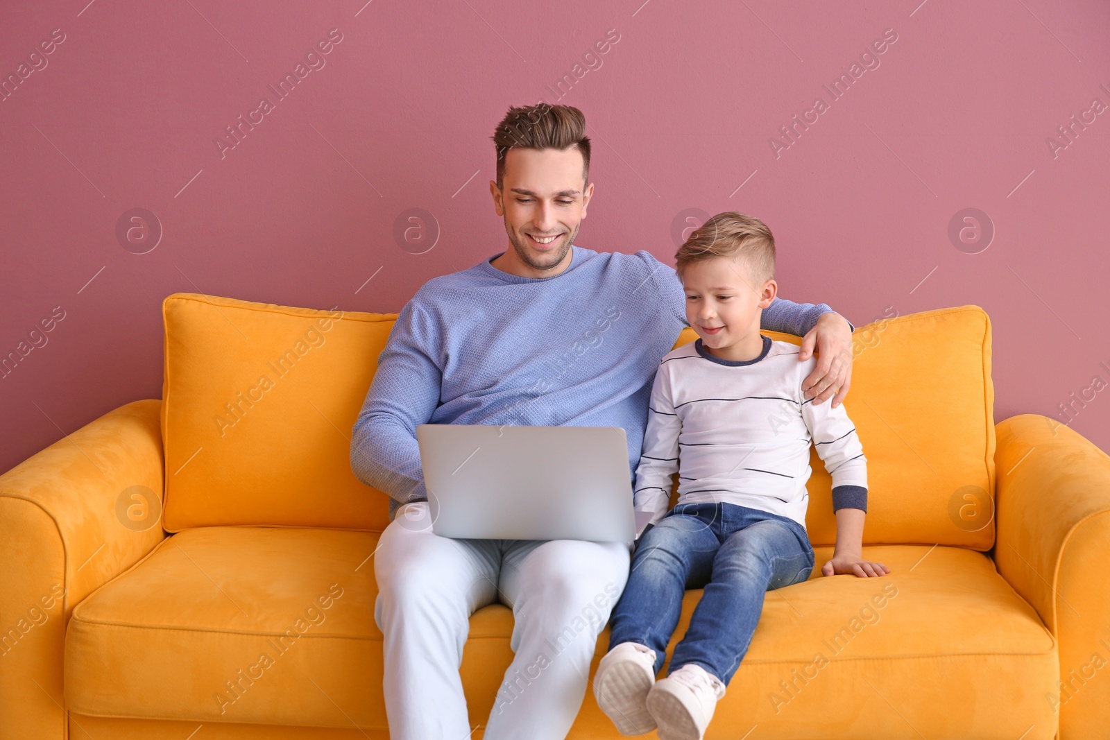 Photo of Little boy and his father with laptop sitting on sofa, indoors