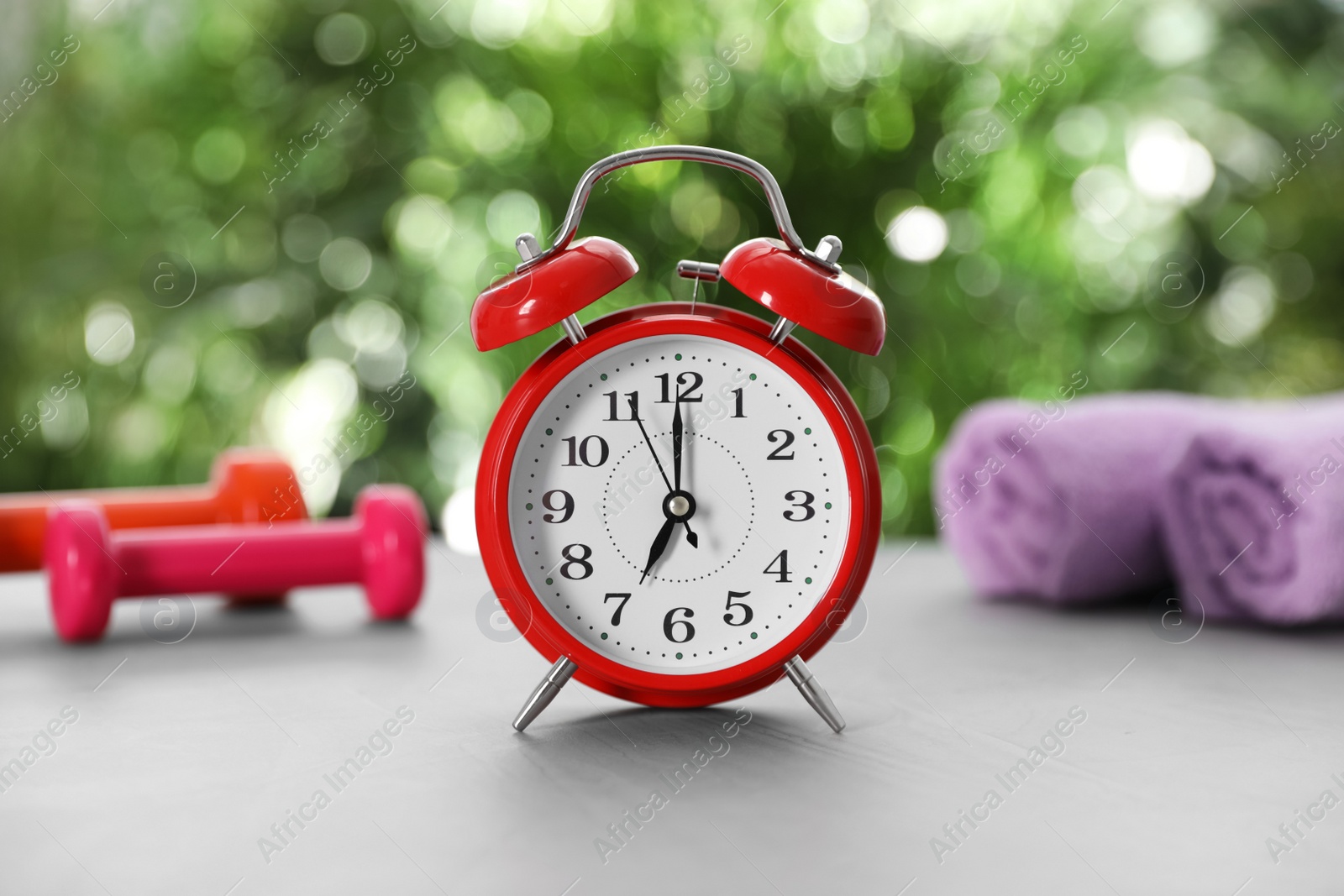 Photo of Alarm clock, towels and dumbbells on grey table against blurred background. Morning exercise