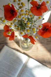 Beautiful bouquet of poppies and chamomiles near open book indoors, focus on flowers