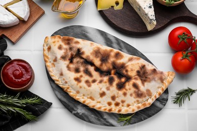 Tasty pizza calzone with tomato sauce and different products on white tiled table, flat lay