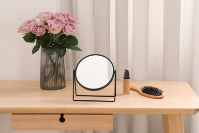 Mirror, foundation, brush and vase with pink roses on wooden dressing table in makeup room