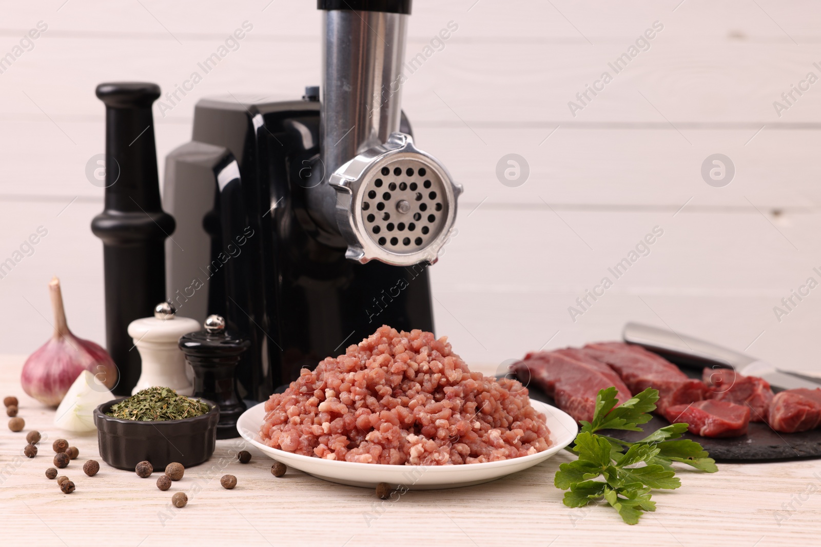 Photo of Electric meat grinder with beef, spices and parsley on white wooden table