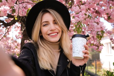 Photo of Happy woman taking selfie with coffee near blossoming sakura outdoors on spring day