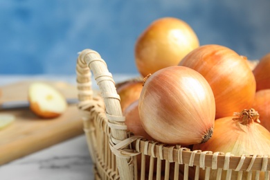Photo of Wicker basket with ripe onions on marble table against blue background, closeup. Space for text