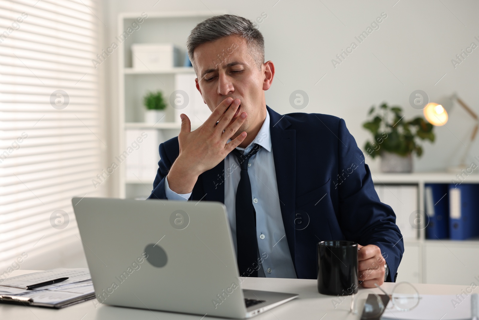 Photo of Sleepy man with cup of drink yawning at table in office