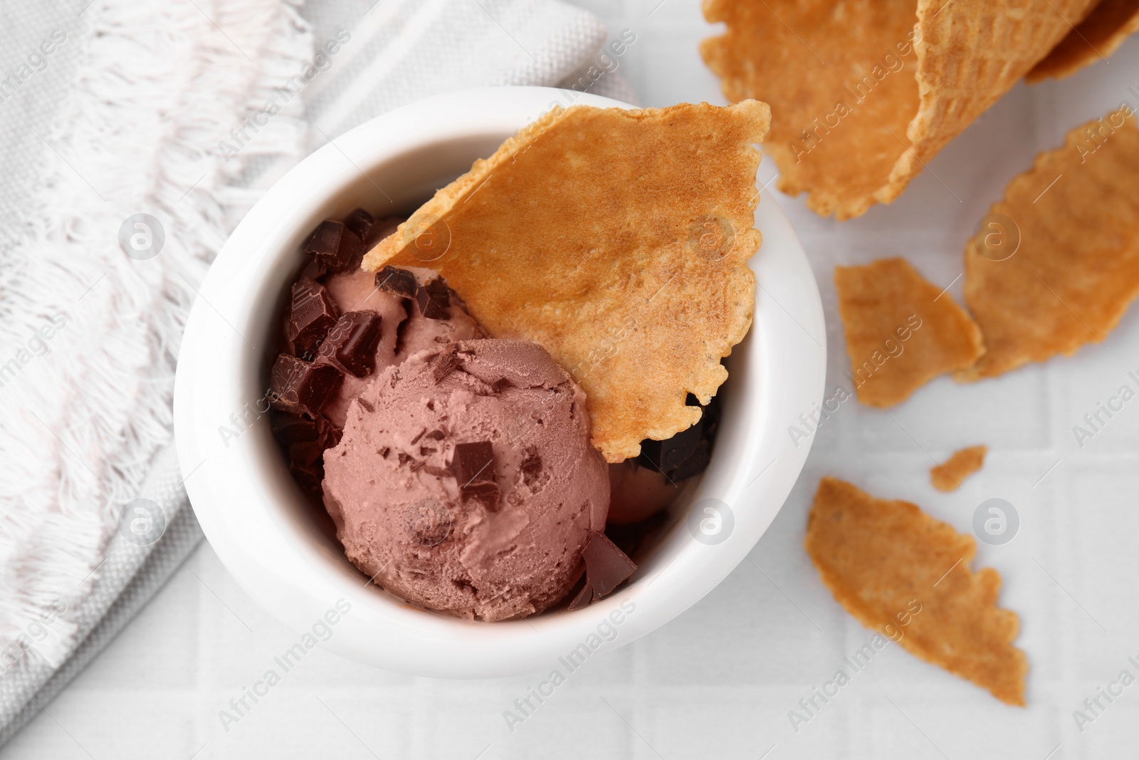 Photo of Tasty chocolate ice cream and pieces of waffle cone in bowl on white table, flat lay