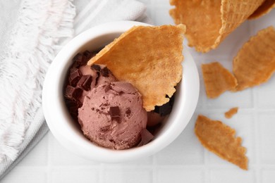 Tasty chocolate ice cream and pieces of waffle cone in bowl on white table, flat lay