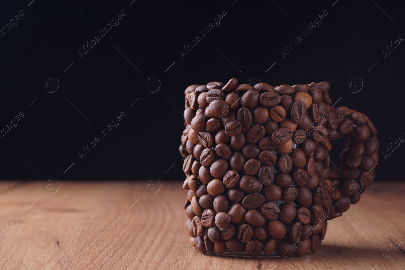Photo of Cup made of coffee beans on wooden table against black background. Space for text