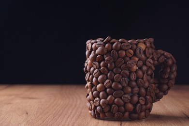 Cup made of coffee beans on wooden table against black background. Space for text