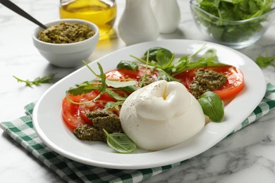 Photo of Delicious burrata salad with tomatoes, arugula and pesto sauce served on white marble table, closeup