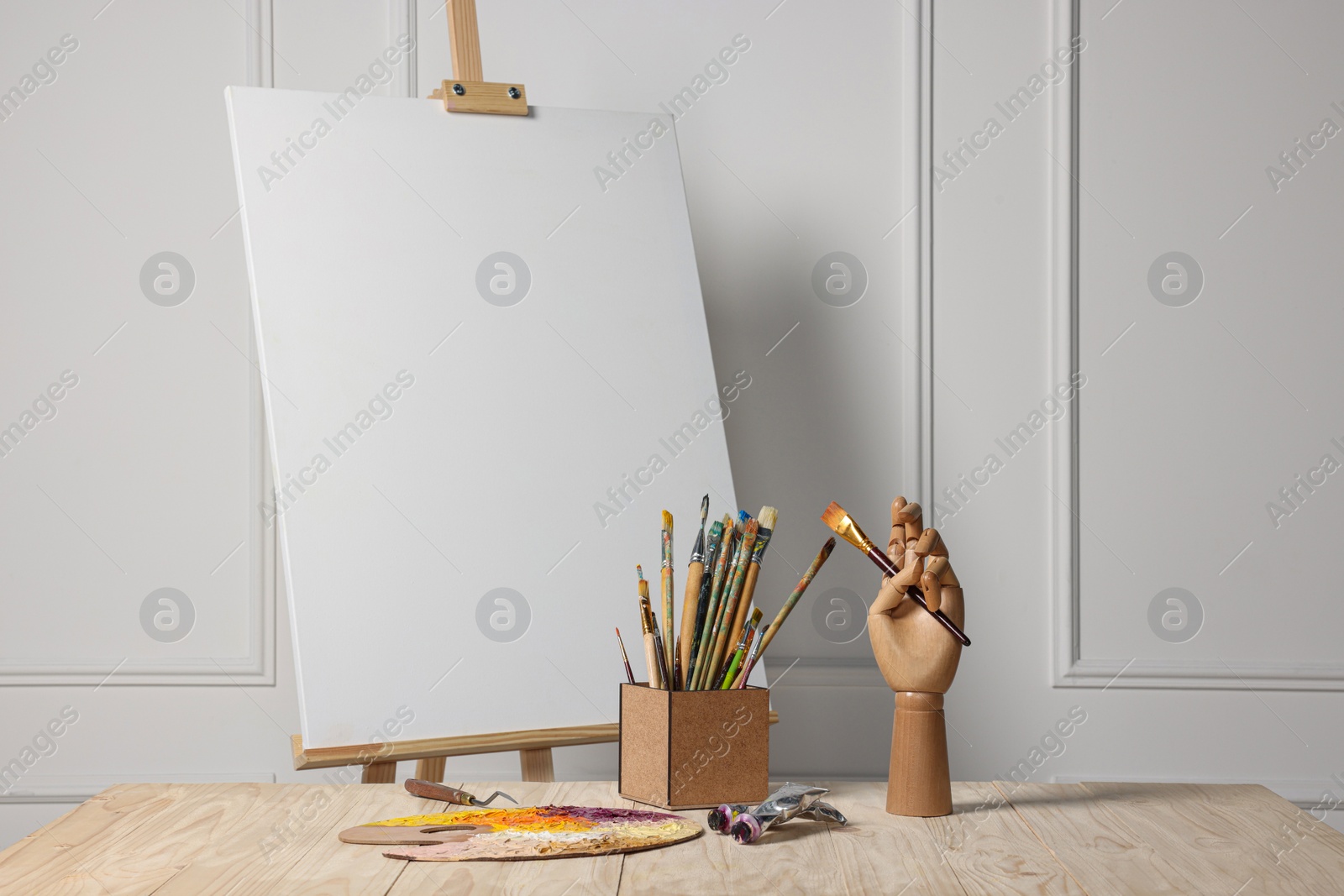 Photo of Easel with blank canvas, hand model and different art supplies on wooden table near white wall