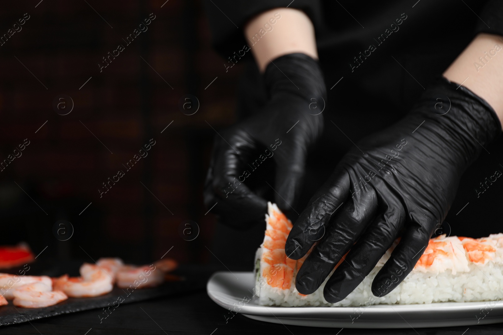 Photo of Chef in gloves making sushi rolls with shrimps at black wooden table, closeup