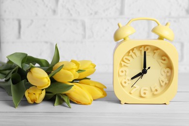 Yellow alarm clock and beautiful tulips on white wooden table against brick wall. Spring time