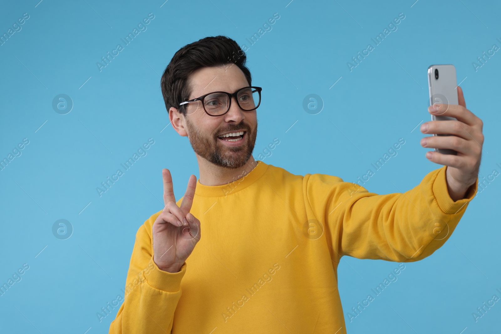 Photo of Smiling man taking selfie with smartphone and showing peace sign on light blue background