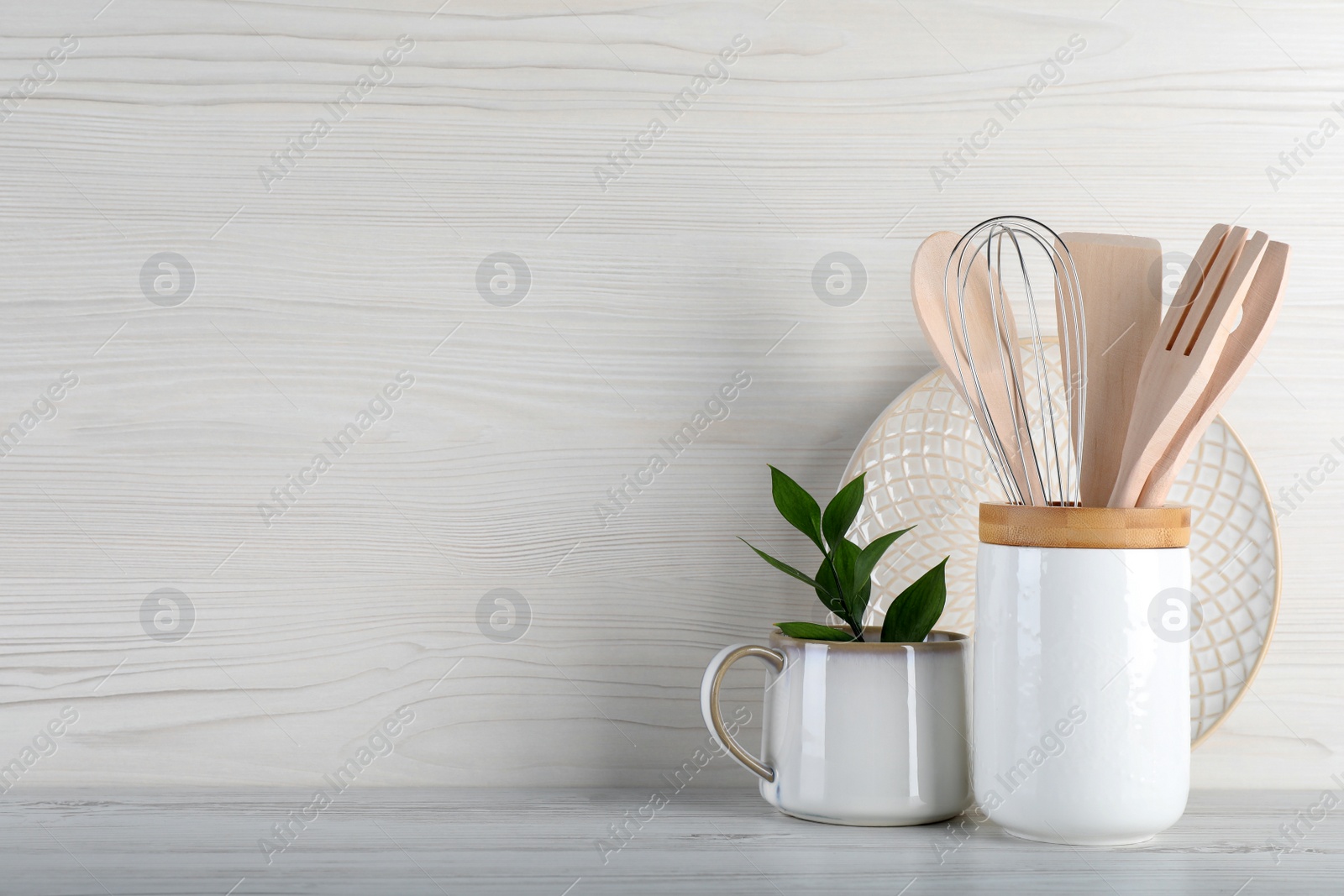 Photo of Holder with kitchen utensils, plate and mug on white wooden table. Space for text