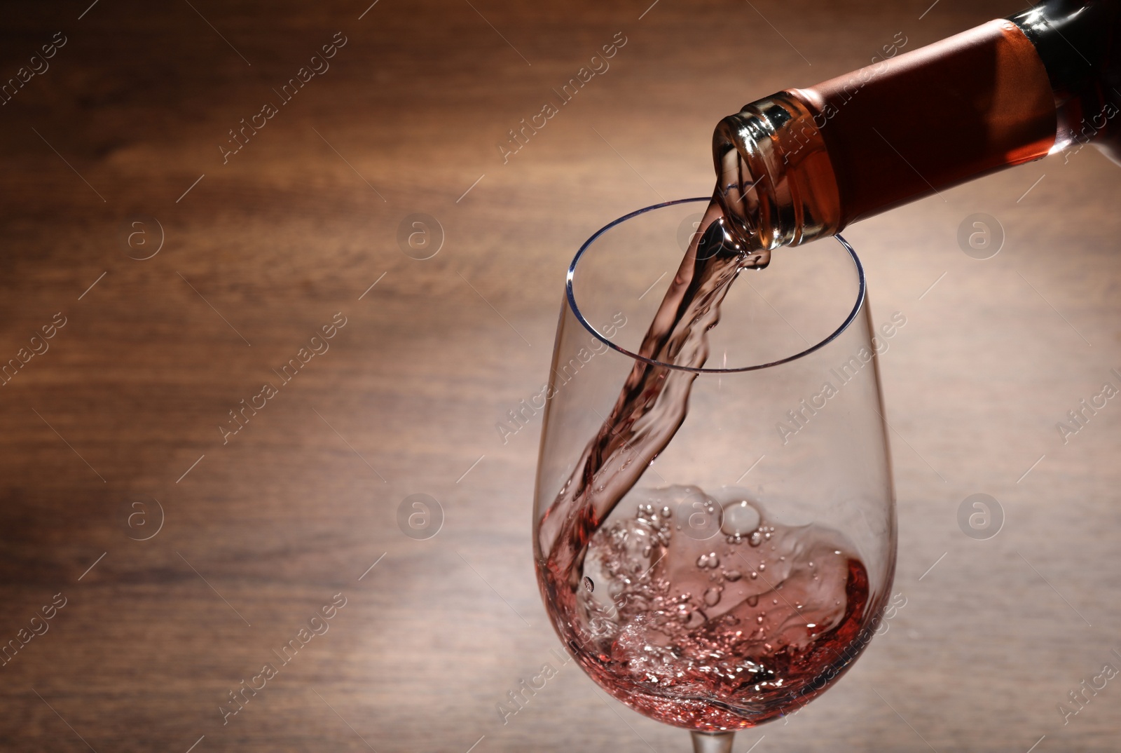 Photo of Pouring rose wine from bottle into glass on wooden table, closeup. Space for text