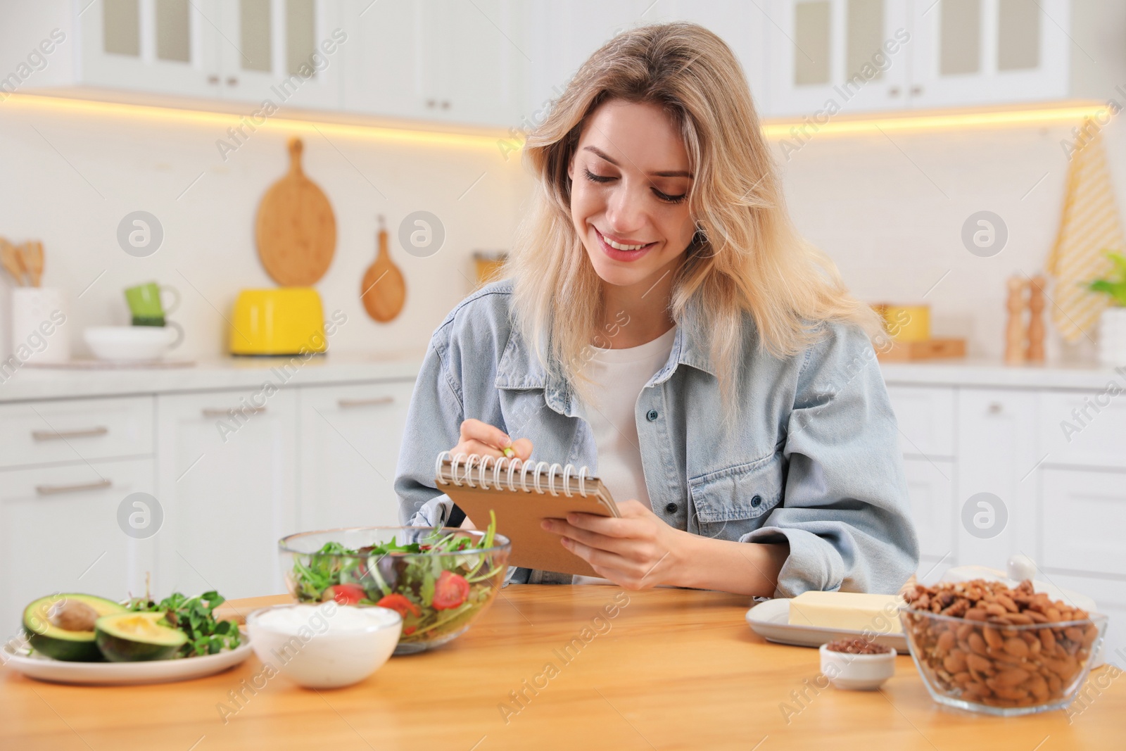 Photo of Woman with notebook and different products at wooden table in kitchen. Keto diet