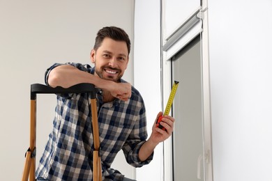 Photo of Man with measure tape on stepladder near window indoors. Roller blinds installation