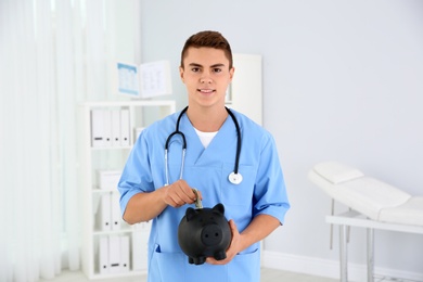Photo of Young medical assistant putting money into piggy bank in clinic