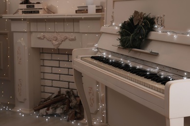 White piano with fairy lights and wreath near decorative fireplace indoors. Christmas music