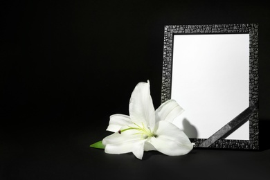 Photo of Funeral photo frame with ribbon and white lily on dark table against black background. Space for design