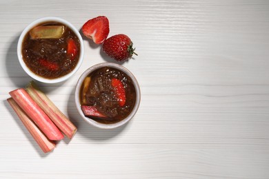 Tasty rhubarb jam in bowls, cut stems and strawberries on white wooden table, flat lay. Space for text