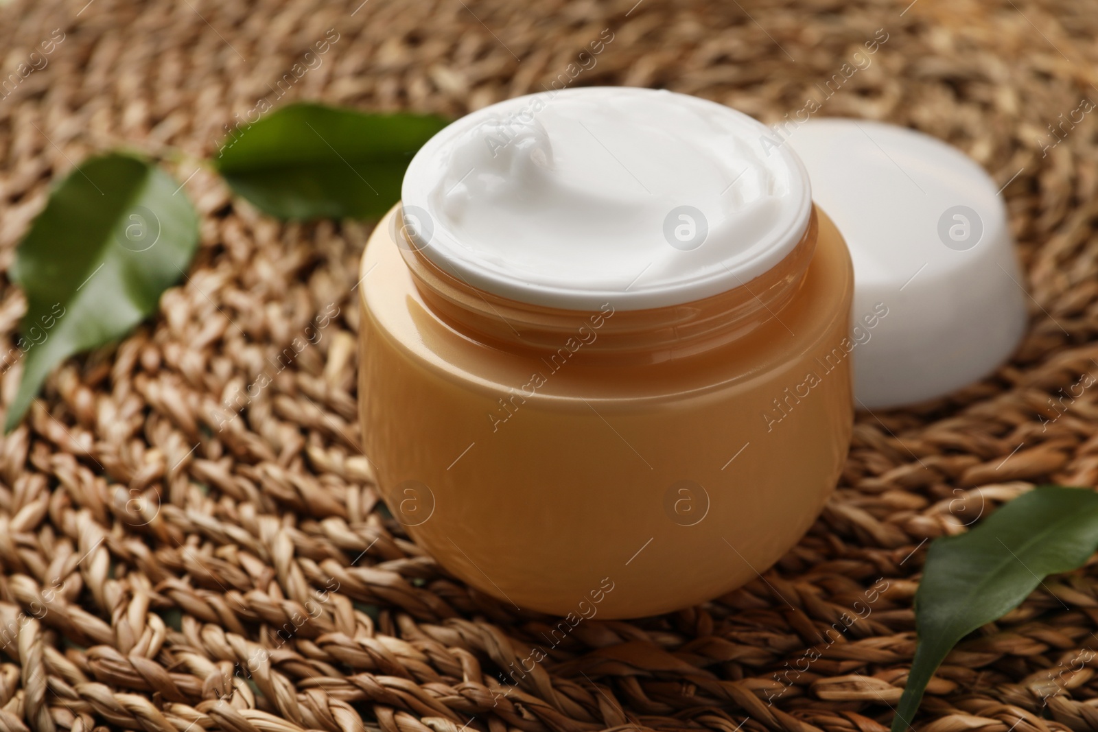 Photo of Jar of face cream and fresh leaves on wicker mat, closeup