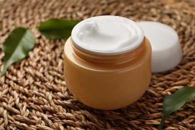 Jar of face cream and fresh leaves on wicker mat, closeup