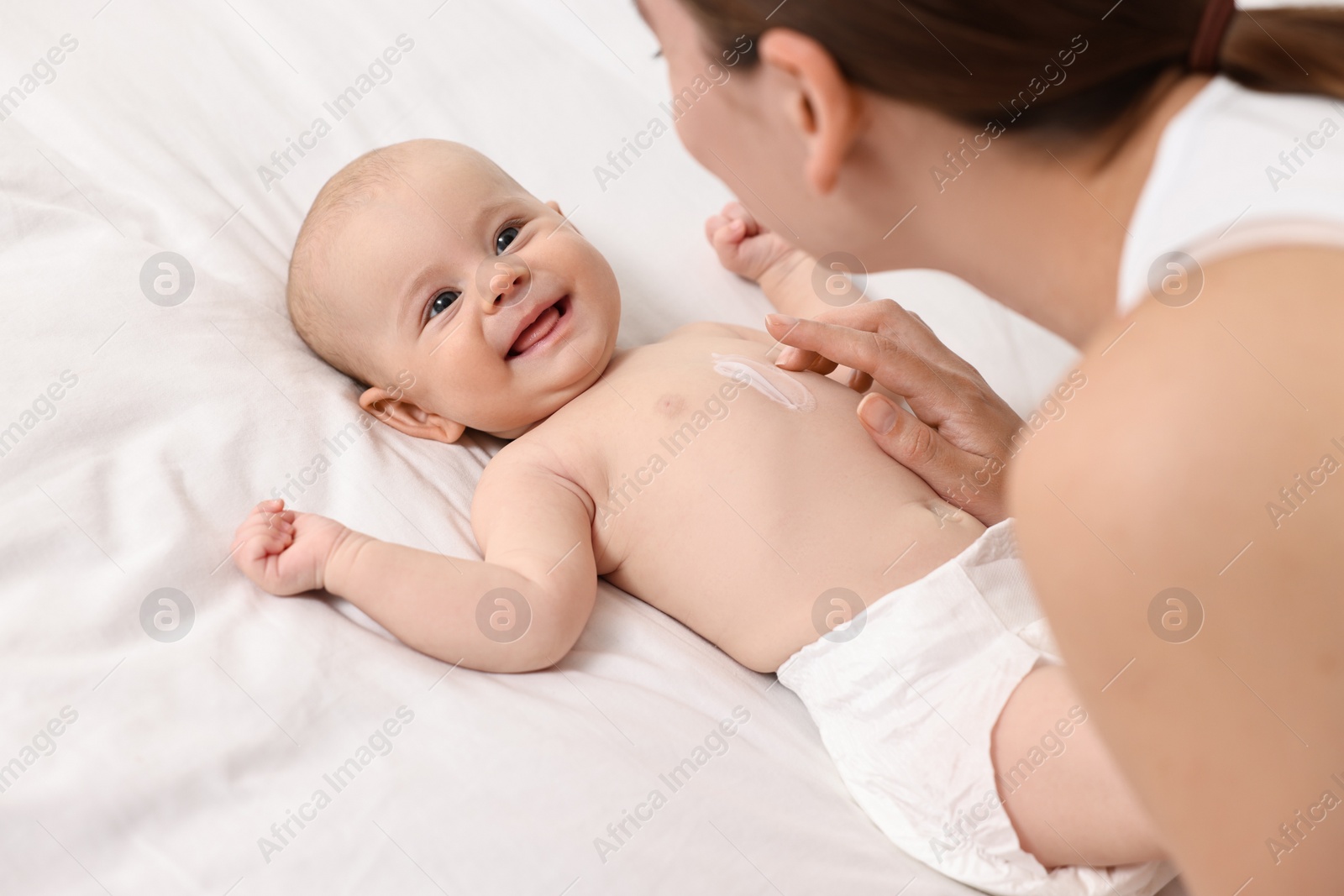 Photo of Woman applying body cream onto baby`s skin on bed, closeup