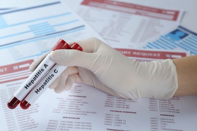 Scientist holding tubes with blood samples for hepatitis virus test against laboratory forms, closeup