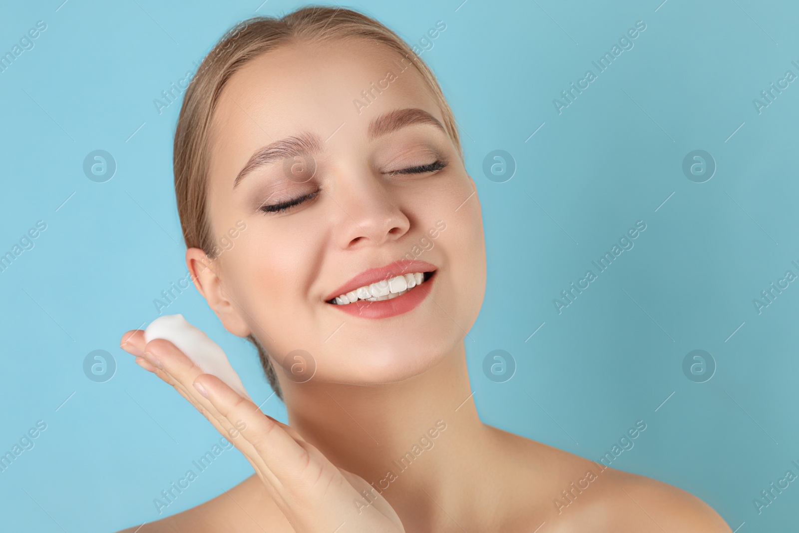 Photo of Young woman washing face with cleansing foam on light blue background. Cosmetic product