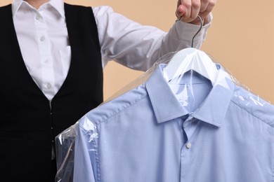 Dry-cleaning service. Woman holding shirt in plastic bag on beige background, closeup