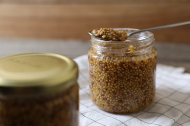Jar and spoon of whole grain mustard on white checkered tablecloth, closeup