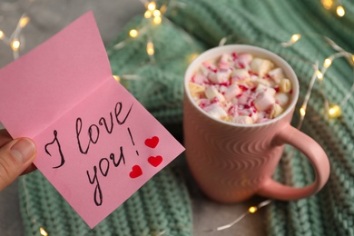 Photo of Woman holding pink note with phrase I Love You near cup of hot drink, closeup