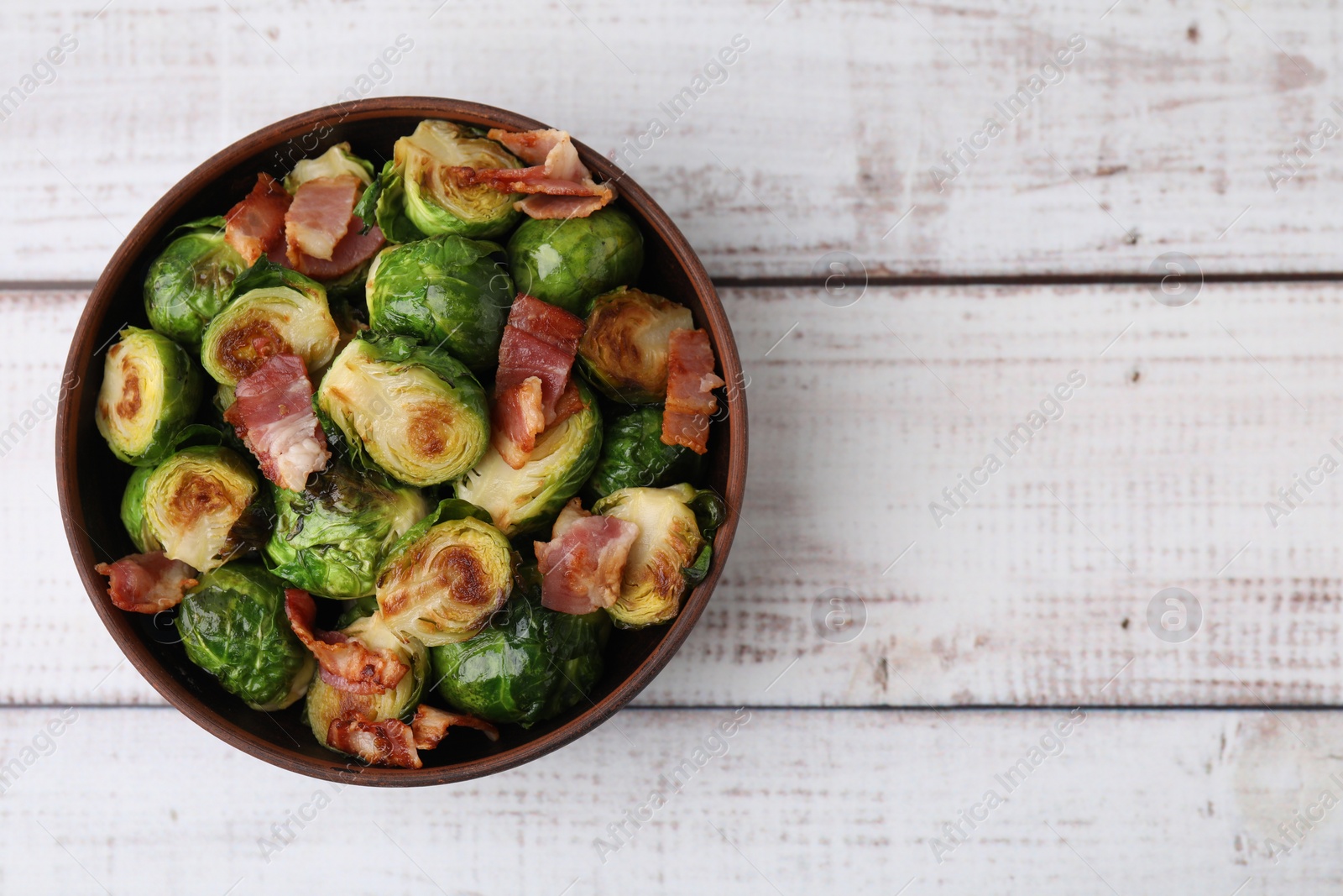 Photo of Delicious roasted Brussels sprouts and bacon in bowl on light wooden table, top view. Space for text