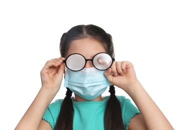 Photo of Little girl wiping foggy glasses caused by wearing medical face mask on white background. Protective measure during coronavirus pandemic