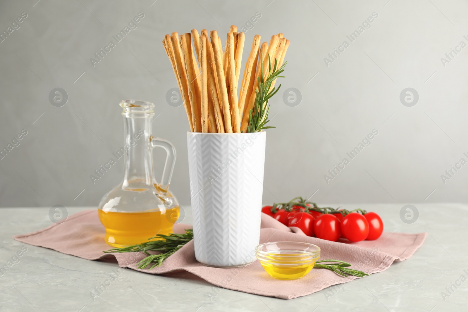 Photo of Delicious grissini sticks, oil, rosemary and tomatoes on grey marble table