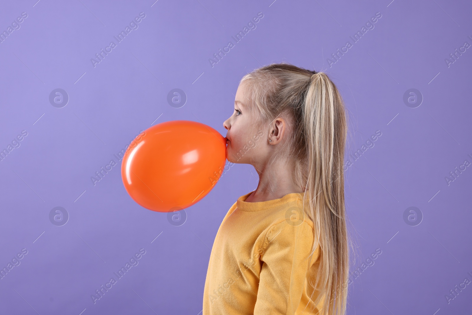 Photo of Cute little girl inflating orange balloon on violet background