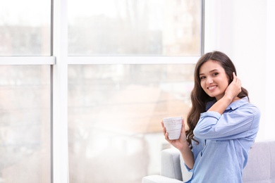 Beautiful young woman with cup of coffee near window