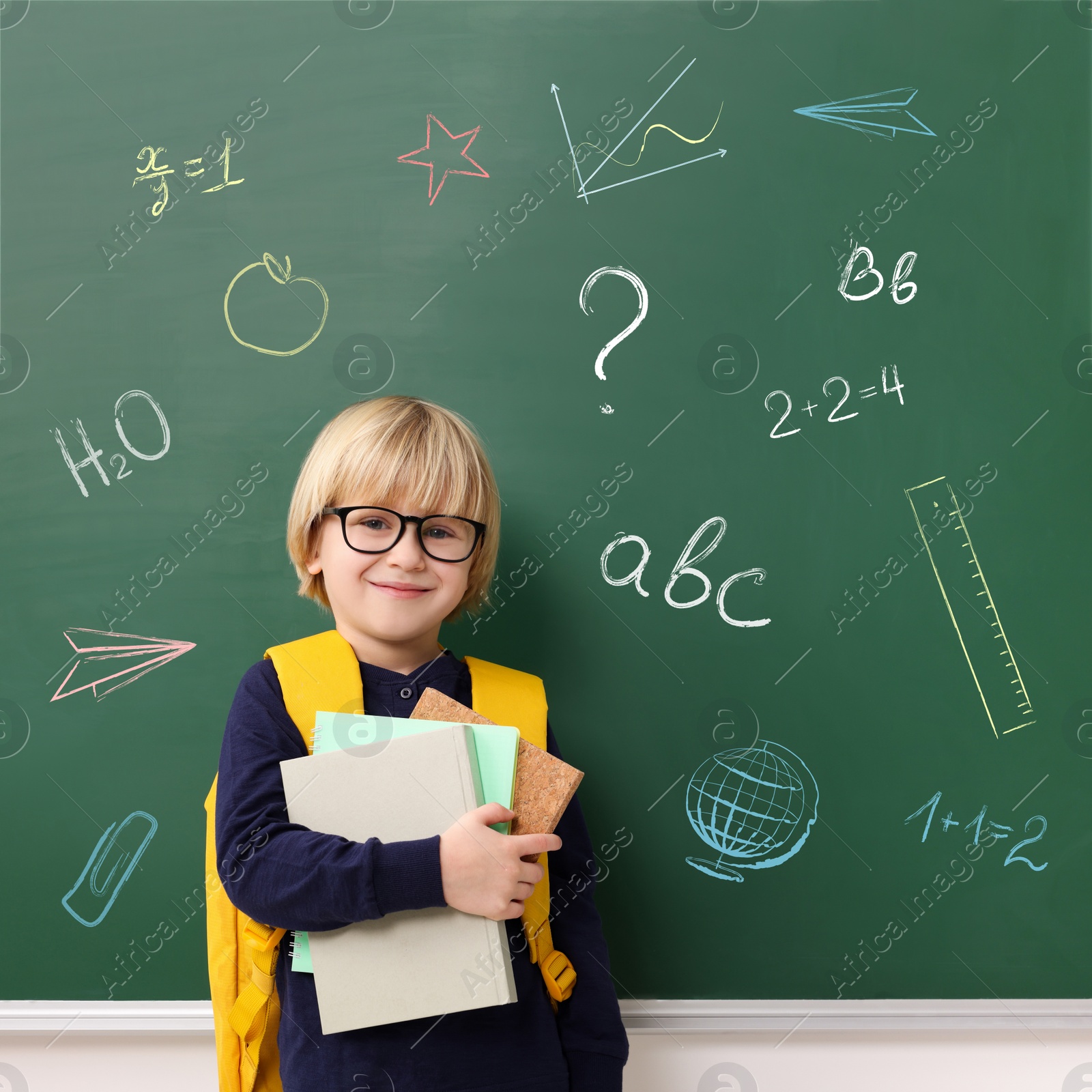 Image of School boy holding notebooks near green chalkboard with drawings and inscriptions