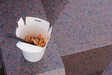 Photo of Paper box of takeaway noodles with fork on stone surface, above view and space for text. Street food