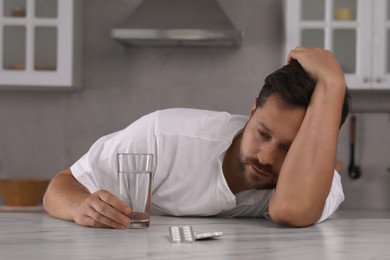 Photo of Depressed man with glass of water and antidepressant pills at table in kitchen