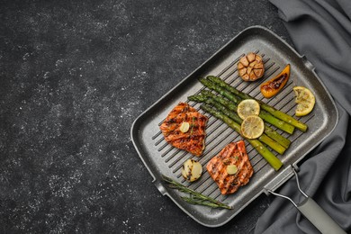 Frying pan with tasty grilled salmon, lemon and asparagus on black table, flat lay. Space for text