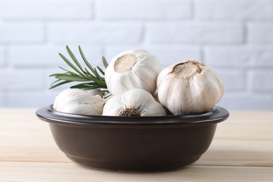 Fresh garlic bulbs in bowl and rosemary on wooden table, closeup