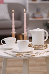 Photo of Cups of tea, teapot and burning candles on white coffee table indoors