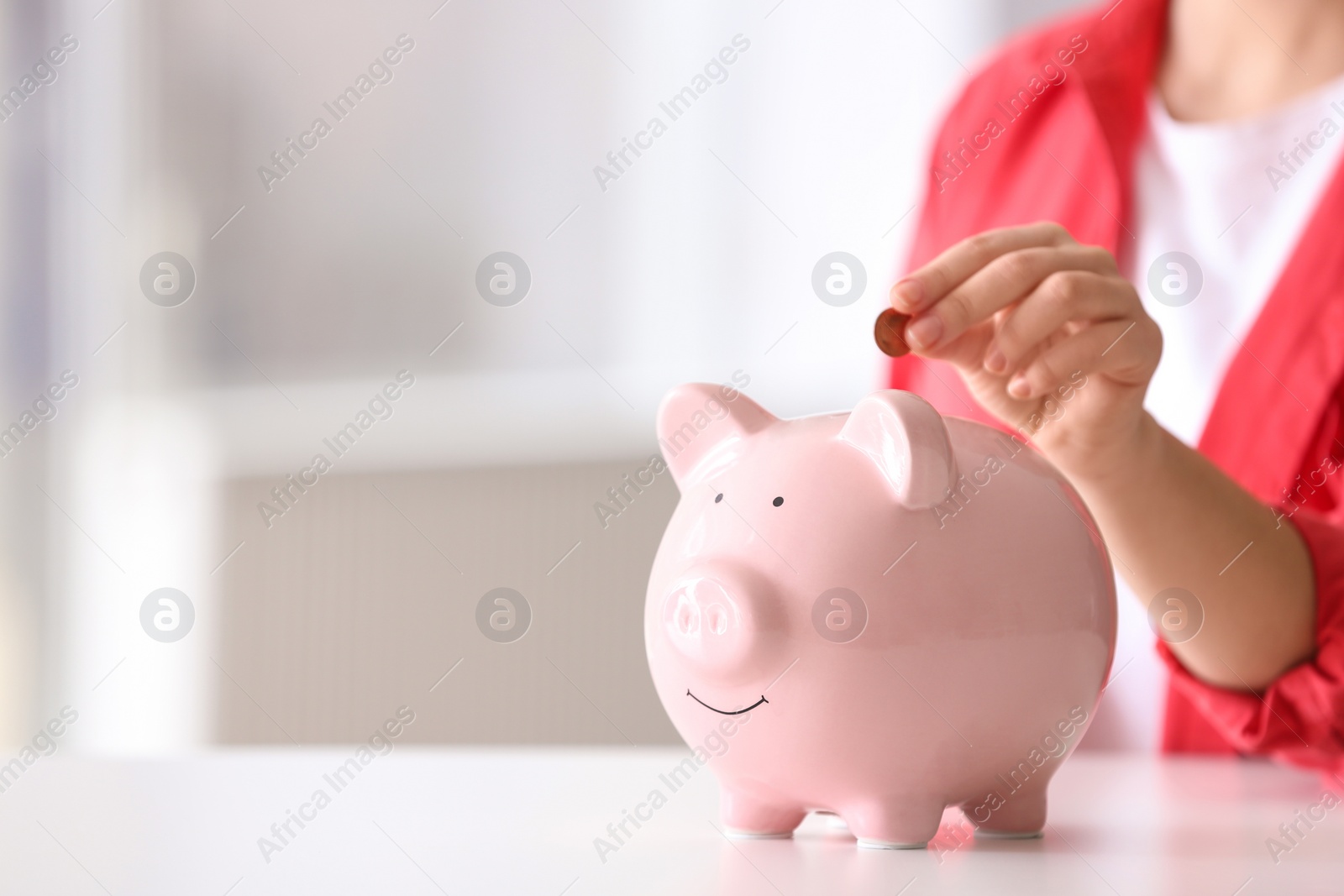 Photo of Woman putting coin into piggy bank at table indoors, closeup. Space for text