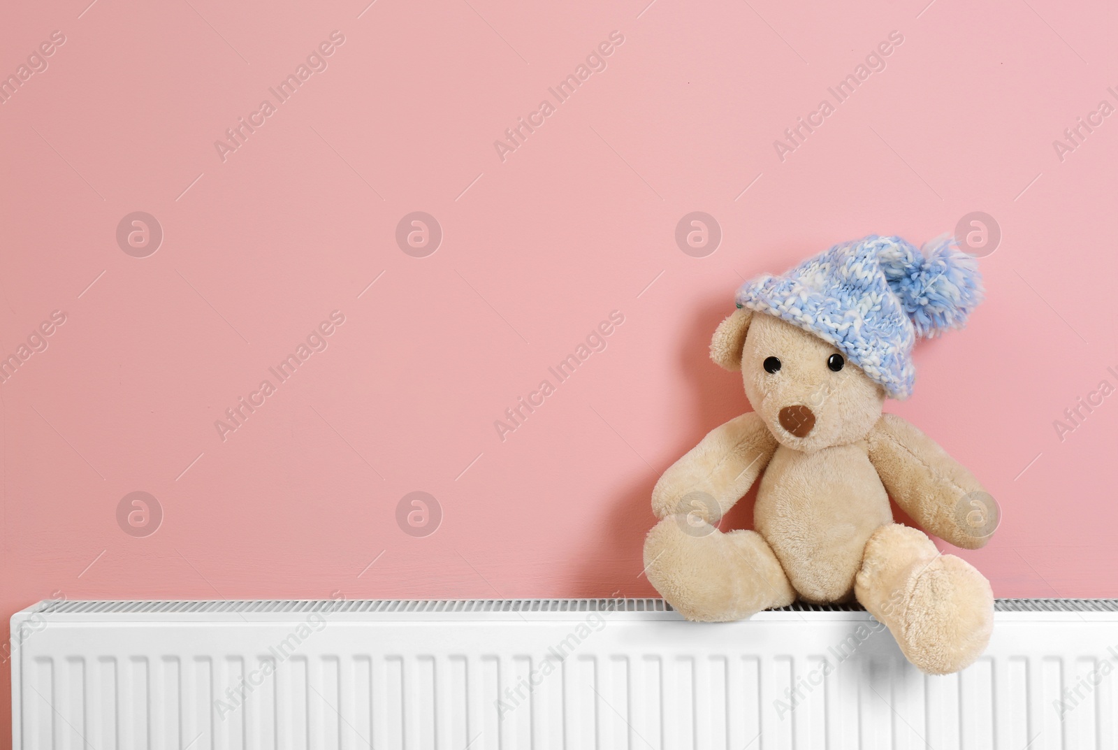 Photo of Teddy bear with knitted hat on heating radiator near color wall. Space for text
