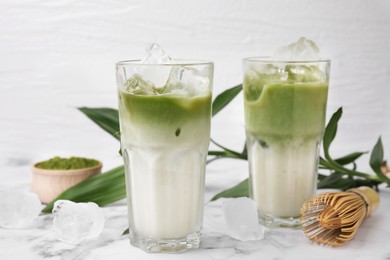 Photo of Glasses of tasty iced matcha latte, bamboo whisk and leaves on white marble table