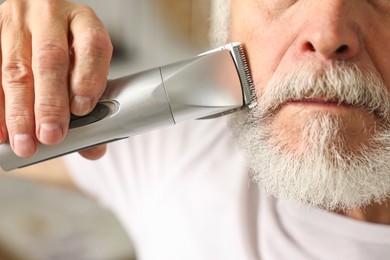 Photo of Man trimming beard with electric trimmer indoors, closeup