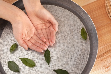 Photo of Woman soaking her hands in bowl with water and leaves on wooden table, top view. Spa treatment
